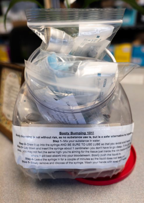Close-up of a clear plastic container with packets of supplies in small bags. A label on the container reads “Booty bumping 101!" with detailed instructions on how to use the supplies