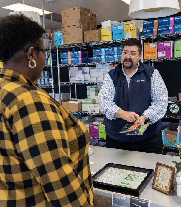 A syringe services program staff person is holding two different-colored packages of syringes with one hand and pointing at one of them with the other while they talk to a client across a counter.